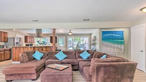 A spacious living room with a large sectional sofa, a kitchen area with wooden cabinets, and a view of the outdoors through large windows. - Family Home w/ Private Pool & Fenced Yard! (Fort Walton Beach)