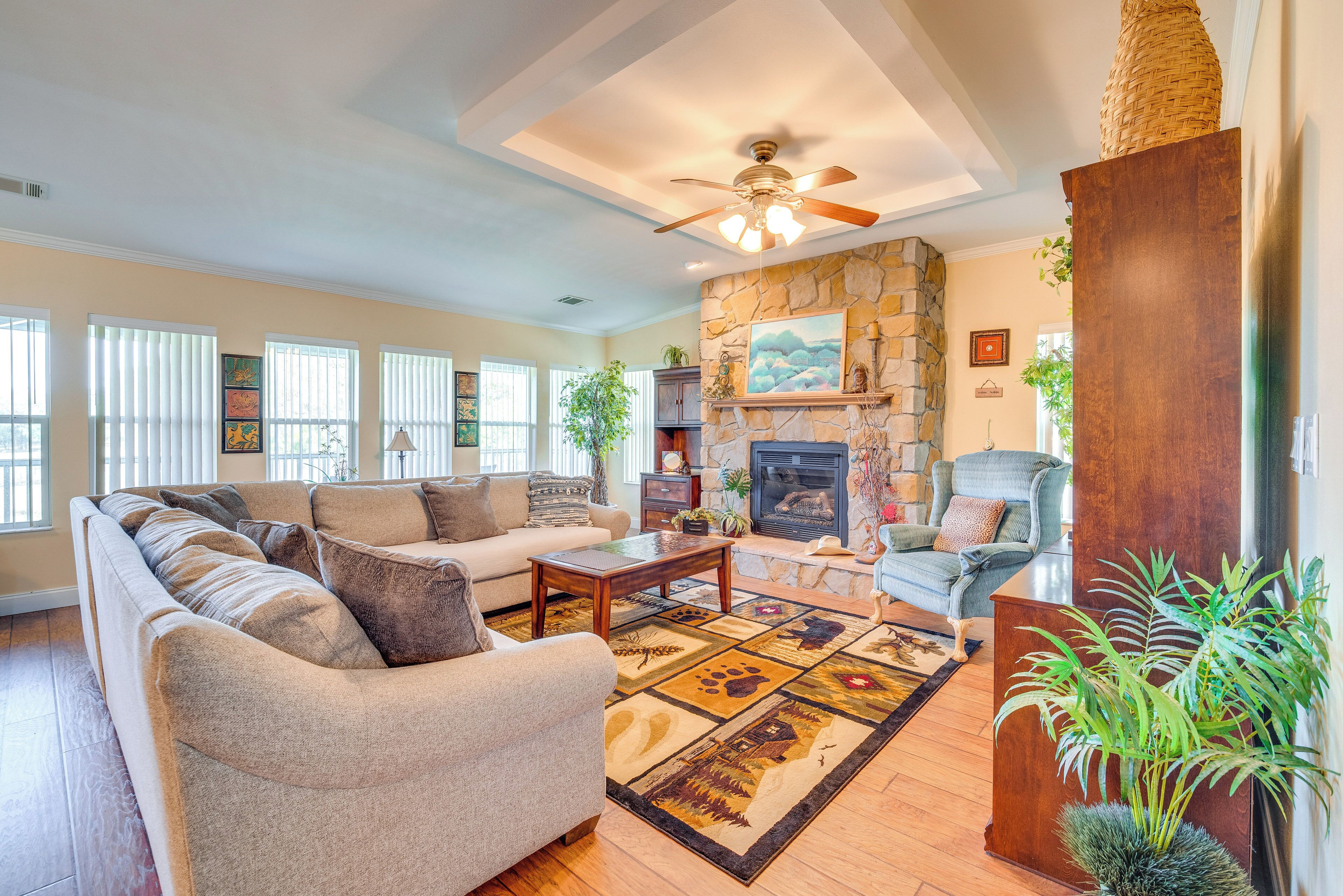 A spacious living room with a stone fireplace, a ceiling fan, and a large window with blinds.