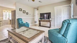 A living room with a glass-top coffee table, a blue armchair, a television on a wooden stand, and a ceiling fan.
