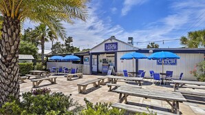 Ein Außenbereich mit Holztischen, Picknicktischen und blauen Stühlen, ein Gebäude mit der Aufschrift "Local Catch Bar & Grill" und Palmen im Hintergrund.