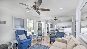 Ein geräumiges Wohnzimmer mit blauem Sessel, beiger Couch und einem gemusterten Teppich. Im Hintergrund befinden sich Deckenventilatoren, ein Essbereich und eine Küche.