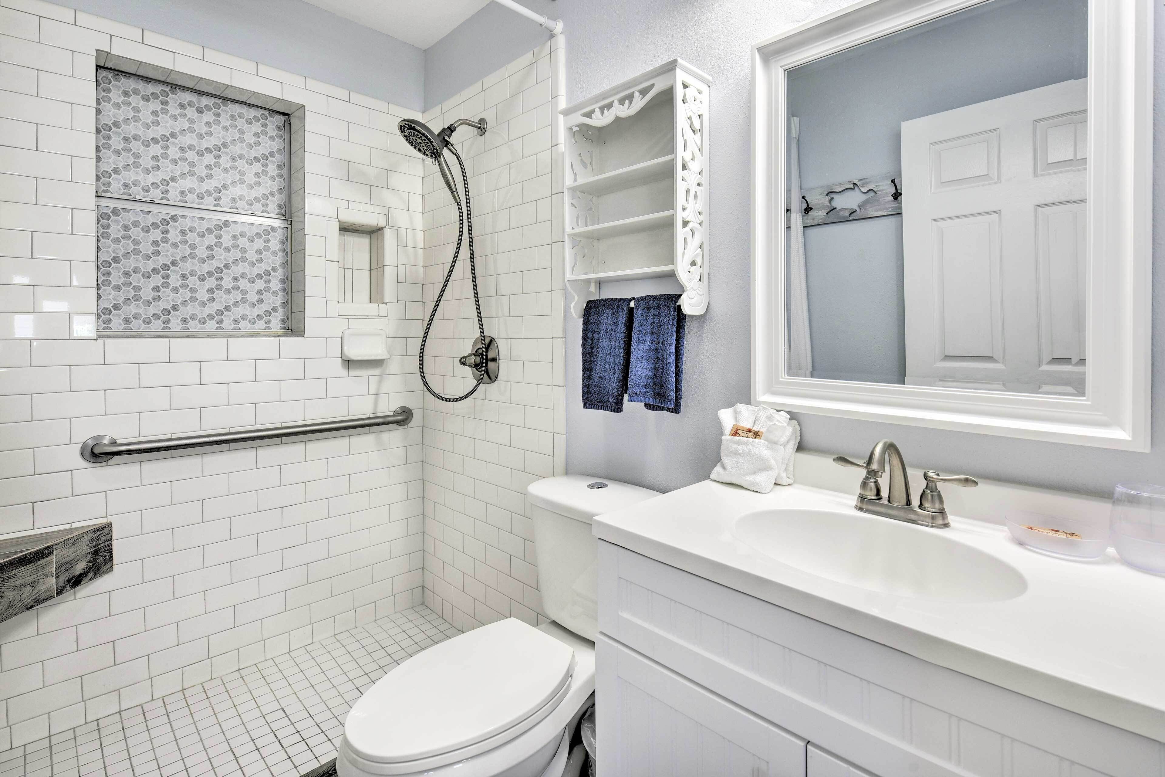 Un baño con ducha de azulejos blancos, inodoro blanco, lavamanos con espejo y toallero blanco.