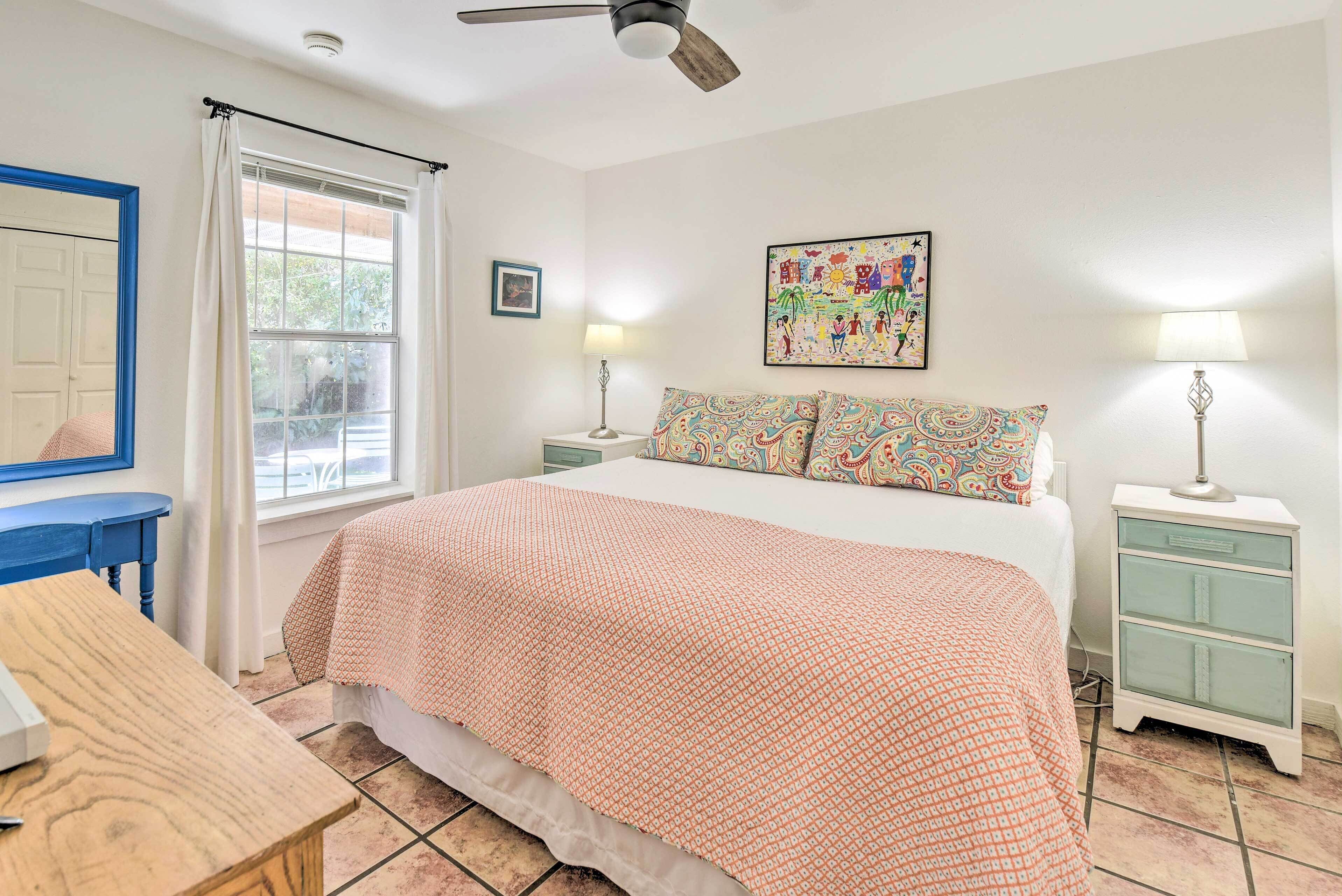 A bedroom with a bed, a desk, a chair, a ceiling fan, a window with curtains, a nightstand, a lamp, and a wall painting.