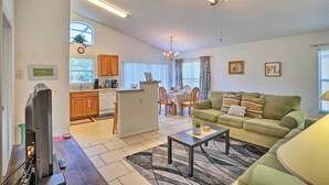 Uma sala de estar moderna com um sofá verde, uma mesa de centro e uma TV. Há uma área de cozinha com armários de madeira e uma mesa de jantar com cadeiras.