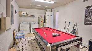 Uma mesa de bilhar com superfície vermelha, uma taça de bilhar e uma bola branca. Ao fundo, uma porta branca com a inscrição "GAME ROOM" e "LADY".