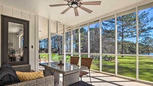 A spacious room with a glass table and wicker furniture, large windows offering a view of a grassy area and trees. - 'green Acres' Home w/ Fire Pit + Fishing Pond (Molino)