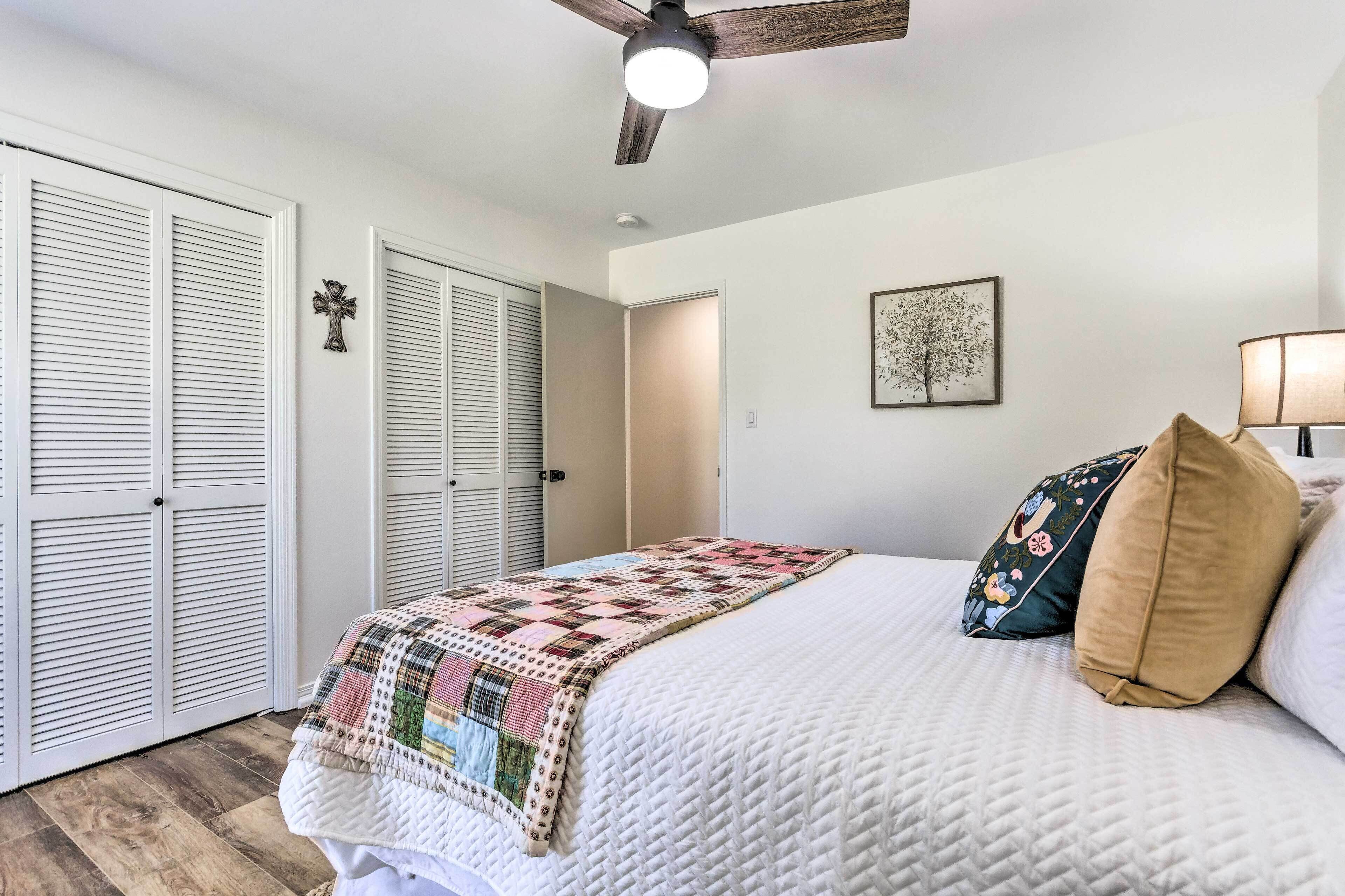 A bedroom with a bed, a quilt, pillows, a ceiling fan, a wall-mounted cross, and a framed picture.