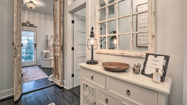 Un couloir avec une porte blanche, un miroir doté d’un cadre rustique, un bol en bois et un panneau encadré posé sur une commode.