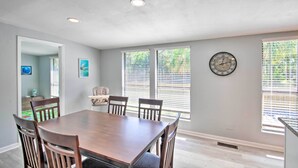 A dining area with a wooden table and chairs, large windows with blinds, a clock on the wall, and a ceiling light.