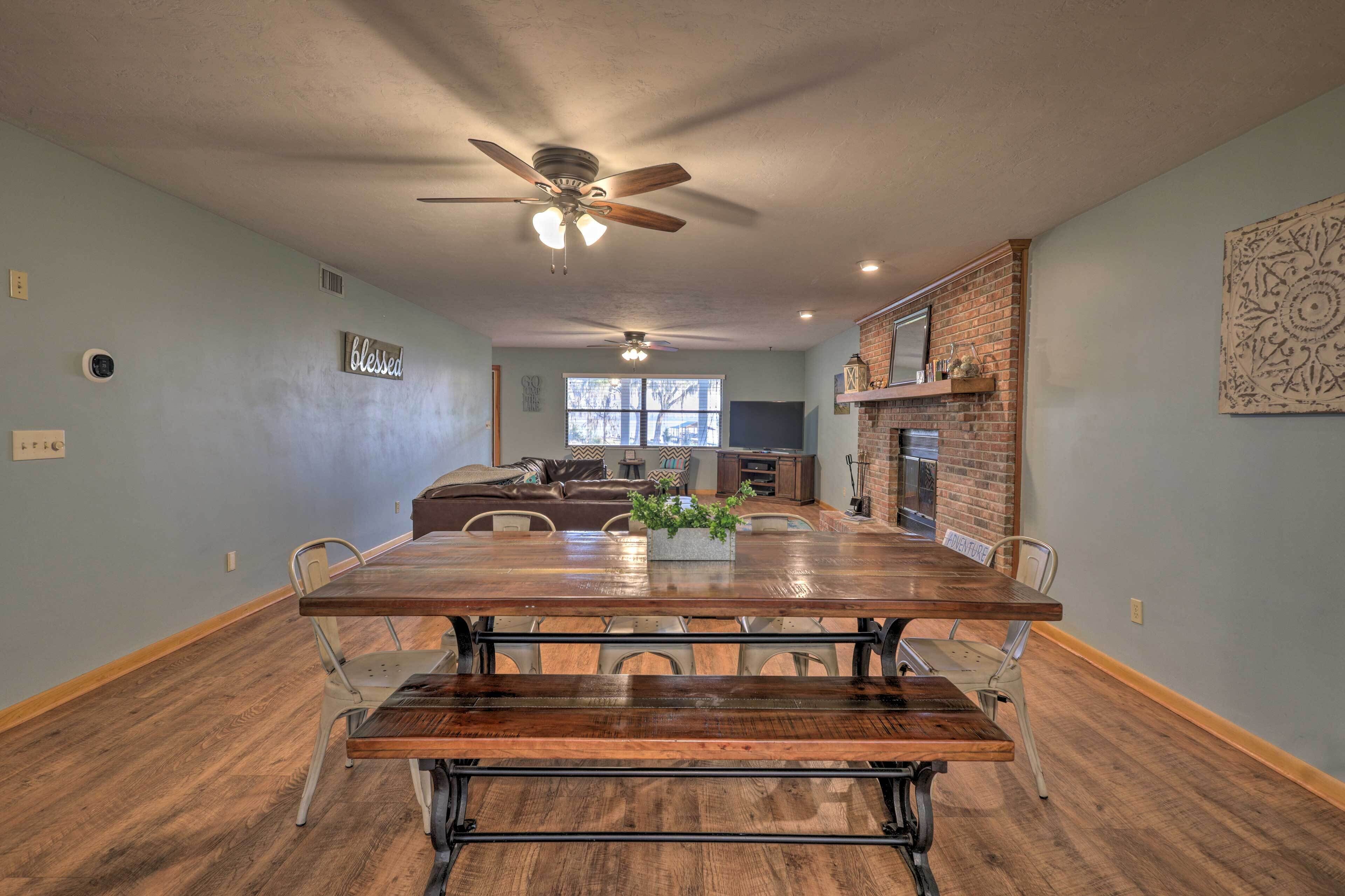 A dining area with a wooden table, metal chairs, and a bench. There is a ceiling fan, a brick fireplace, and a window in the background.