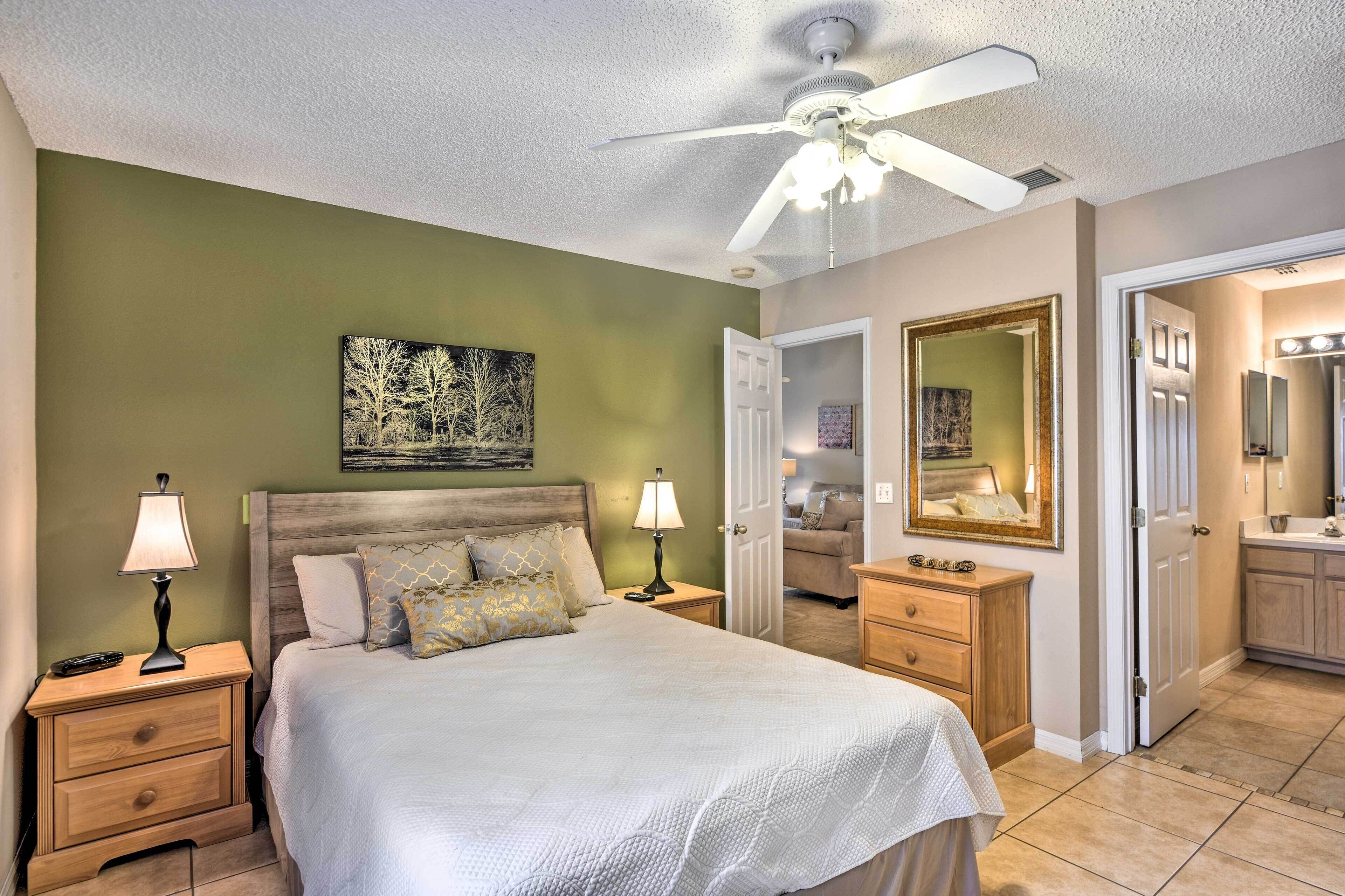 A bedroom with a bed, nightstands, a ceiling fan, a painting, a door leading to a bathroom, and a vanity with a mirror.
