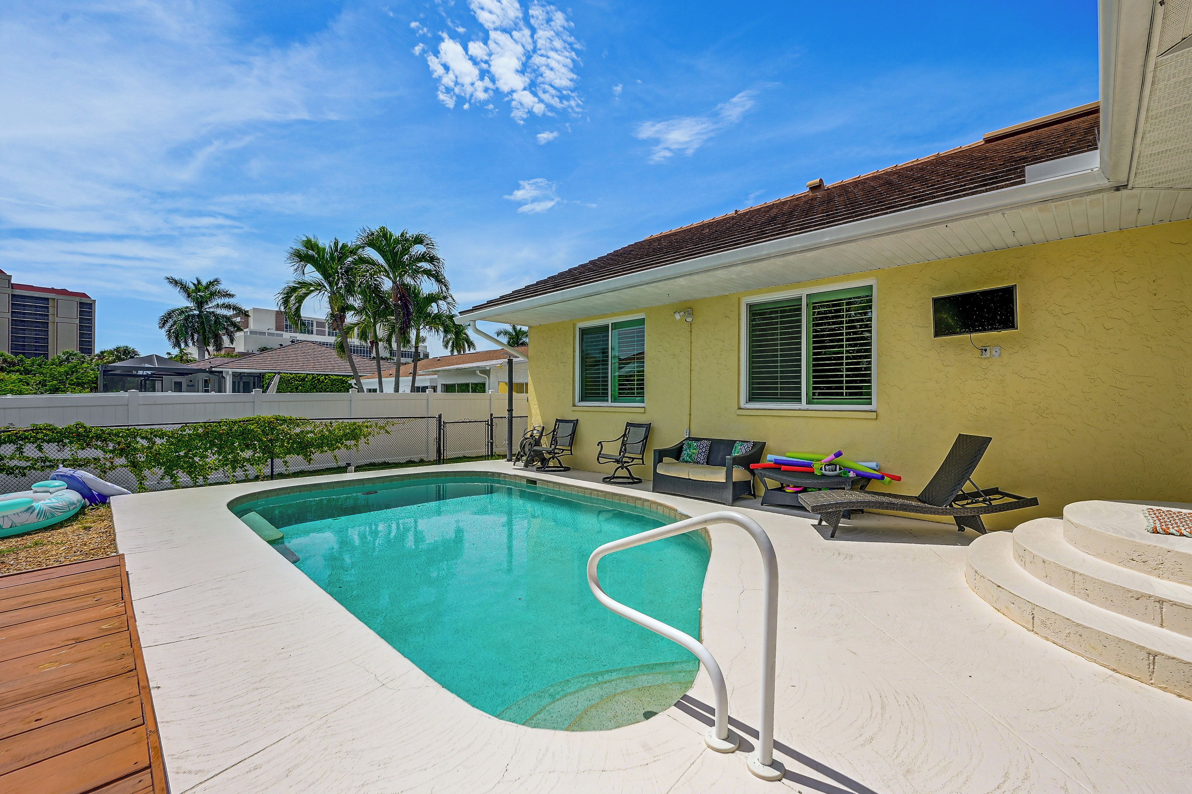 A swimming pool with a wooden deck, lounge chairs, and a yellow building with windows.
