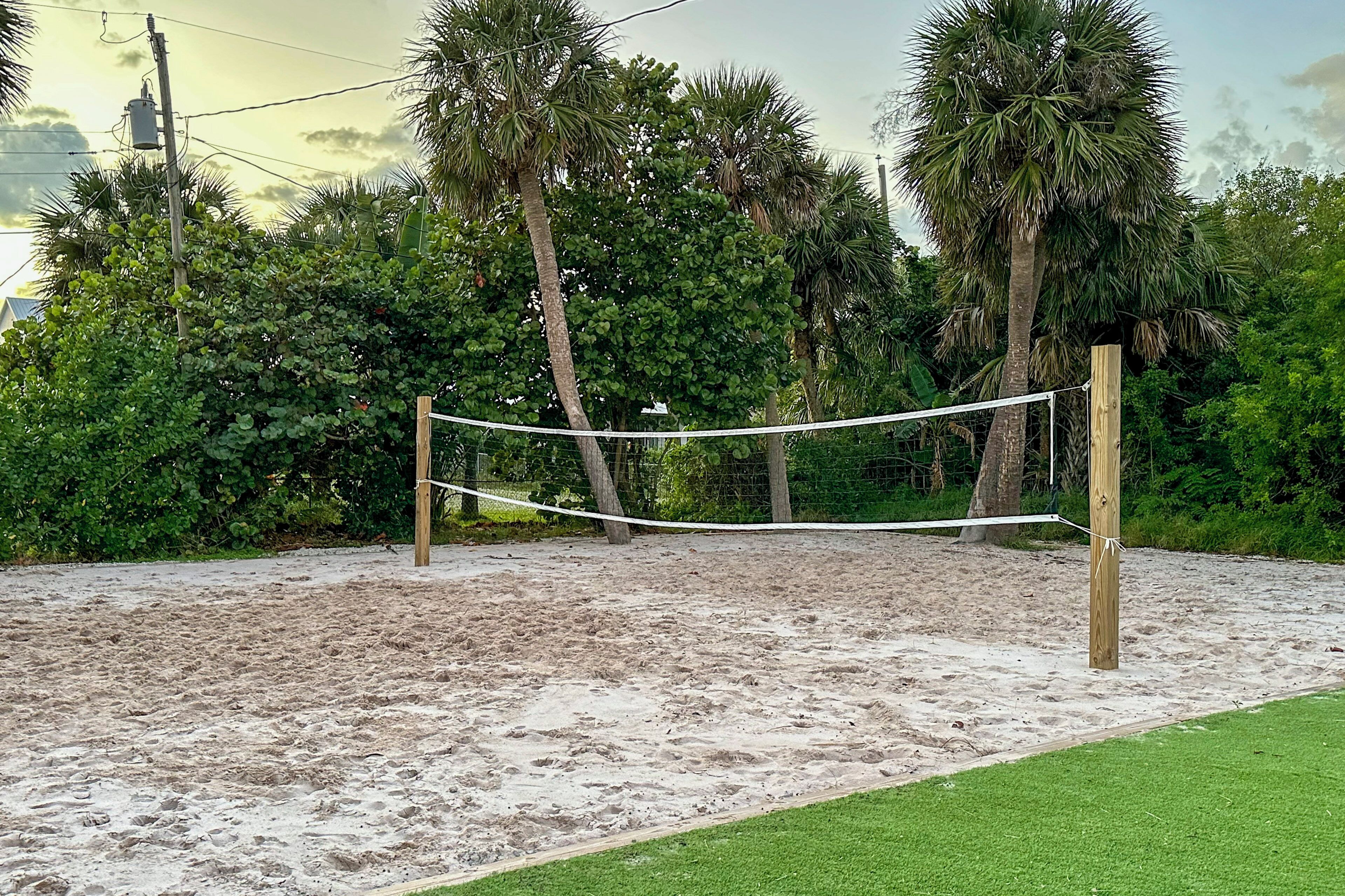 A sandy beach volleyball court with a net, surrounded by palm trees and greenery.