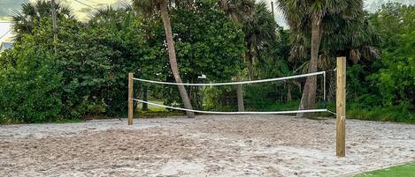 A sandy beach volleyball court with a net, surrounded by palm trees and greenery.