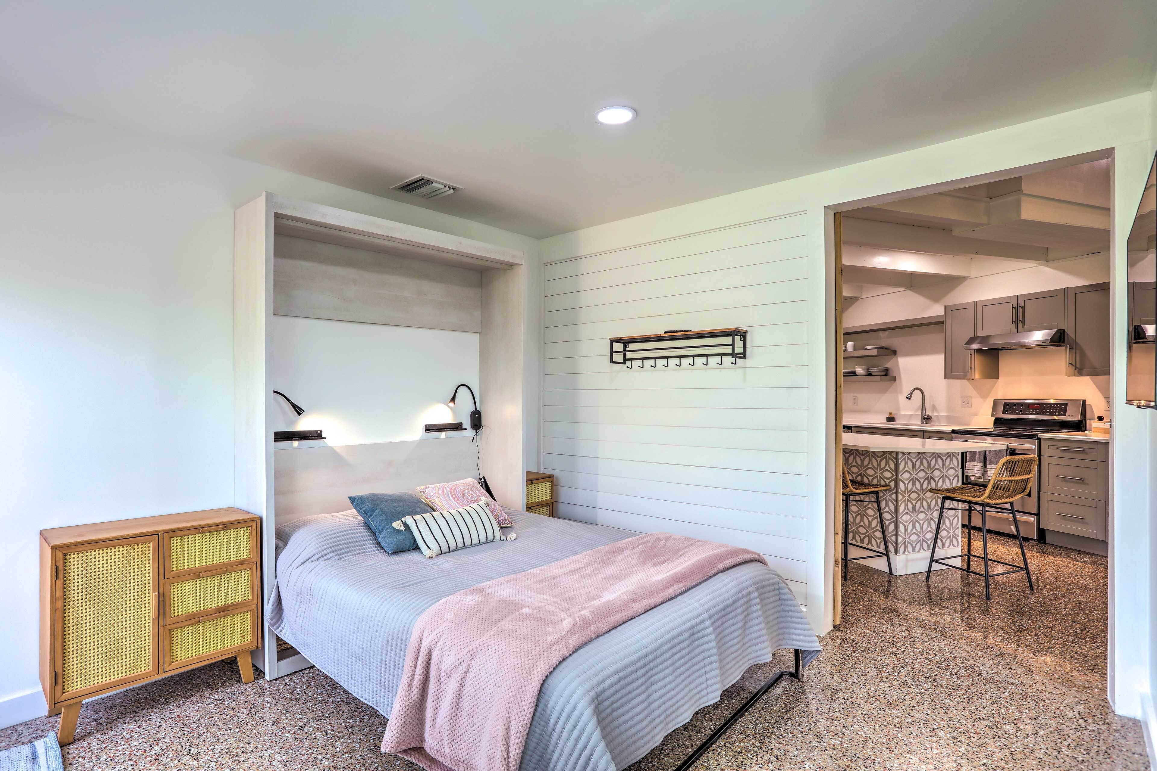 A bedroom with a bed, bedside table, and a small kitchenette area with a sink and bar stools.