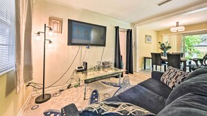 A living room with a flat-screen TV mounted on the wall, a glass coffee table, a sofa with patterned cushions, and a dining area visible through an open door. - Jacksonville Getaway w/ Yard ~ 2 Mi to Beach! (Atlantic Beach)