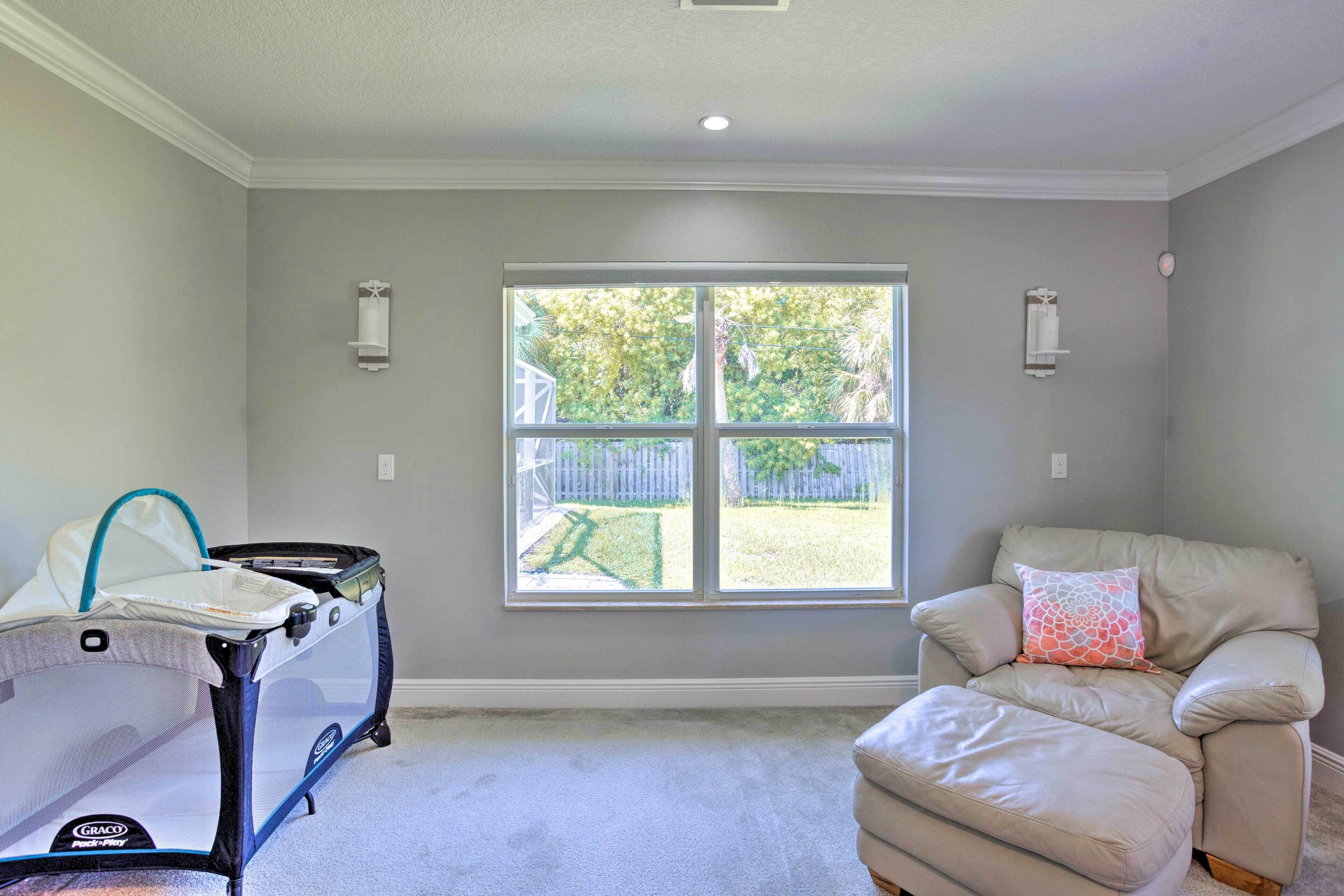 A room with a large window, a baby playpen, and a recliner with a cushion.