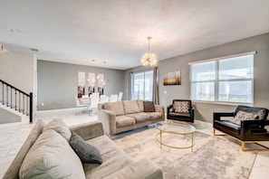A spacious living room with a beige sofa, a black leather sofa, and a glass coffee table. There is a staircase leading to another room, and a large window with blinds.