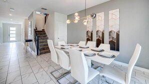 Une salle à manger avec une longue table, des chaises blanches et un lustre. Un escalier avec une rampe mène à une autre pièce.