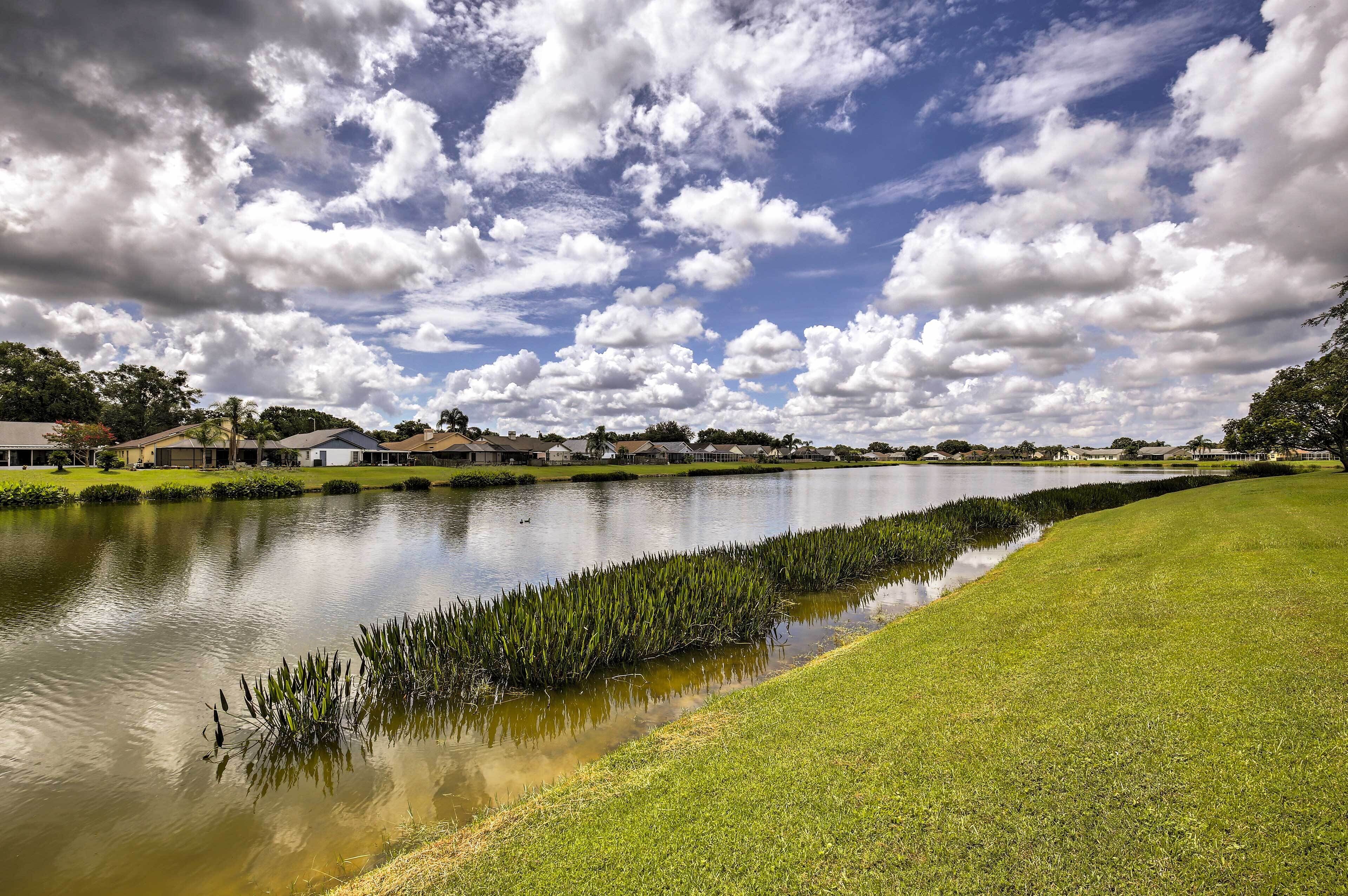 Un lac paisible aux rives verdoyantes, bordé de maisons et d’arbres, sous un ciel partiellement nuageux.