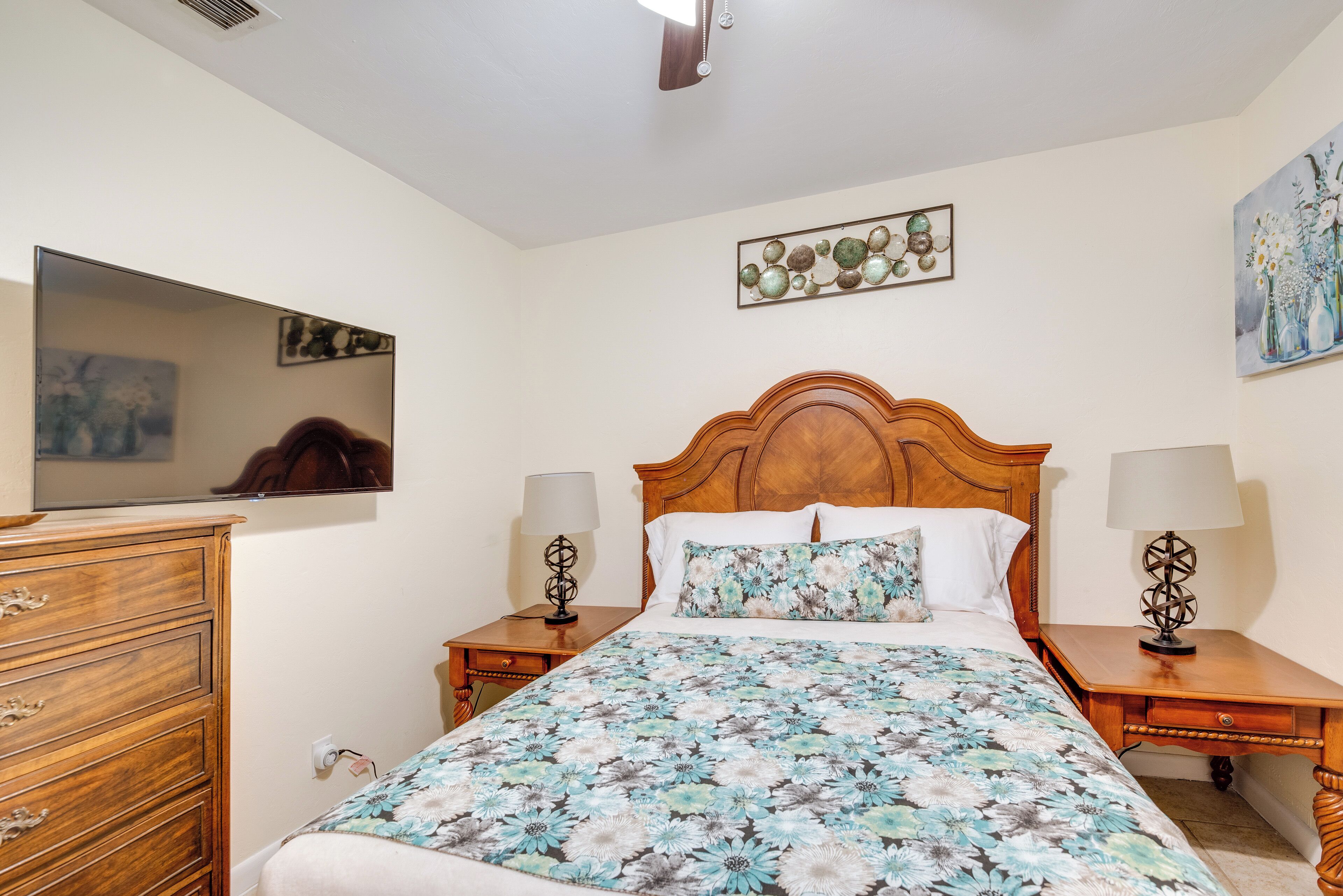 Une chambre à coucher avec une tête de lit en bois, une couvre-lit à motifs floraux, deux tables de chevet avec des lampes, une commode et un téléviseur à écran plat fixé au mur.