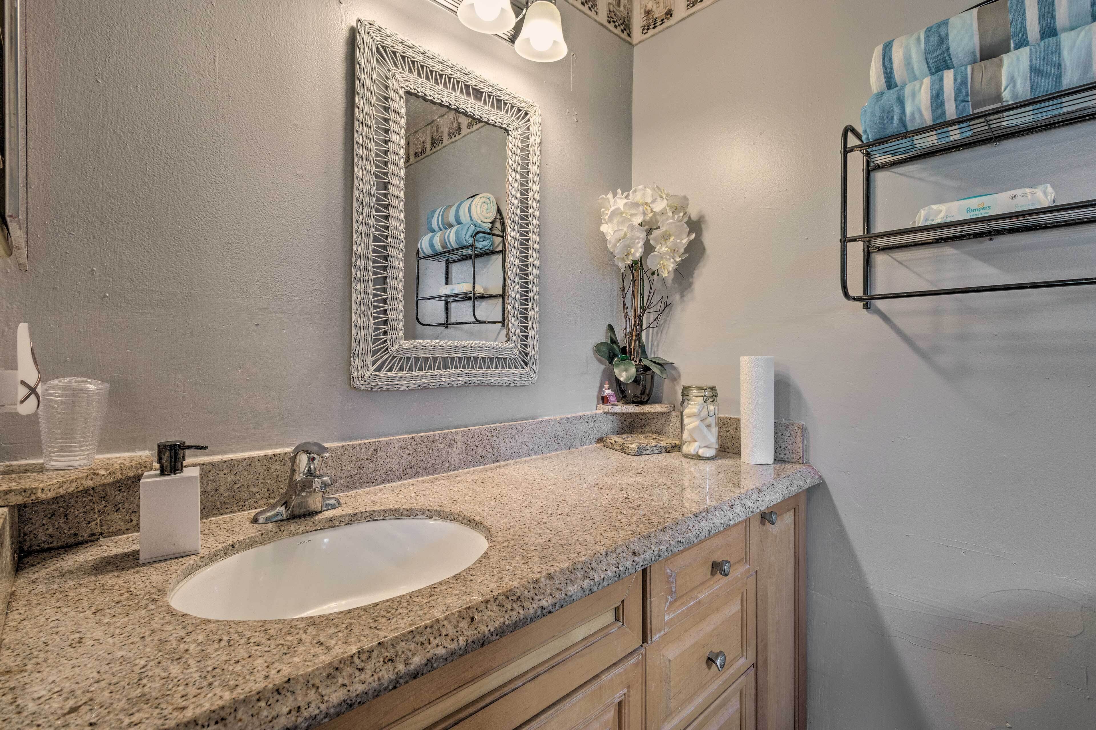 Uma casa de banho com bancada de granito, pia branca, espelho com moldura decorativa, porta-toalhas com toalha listrada e um vaso com flores brancas.