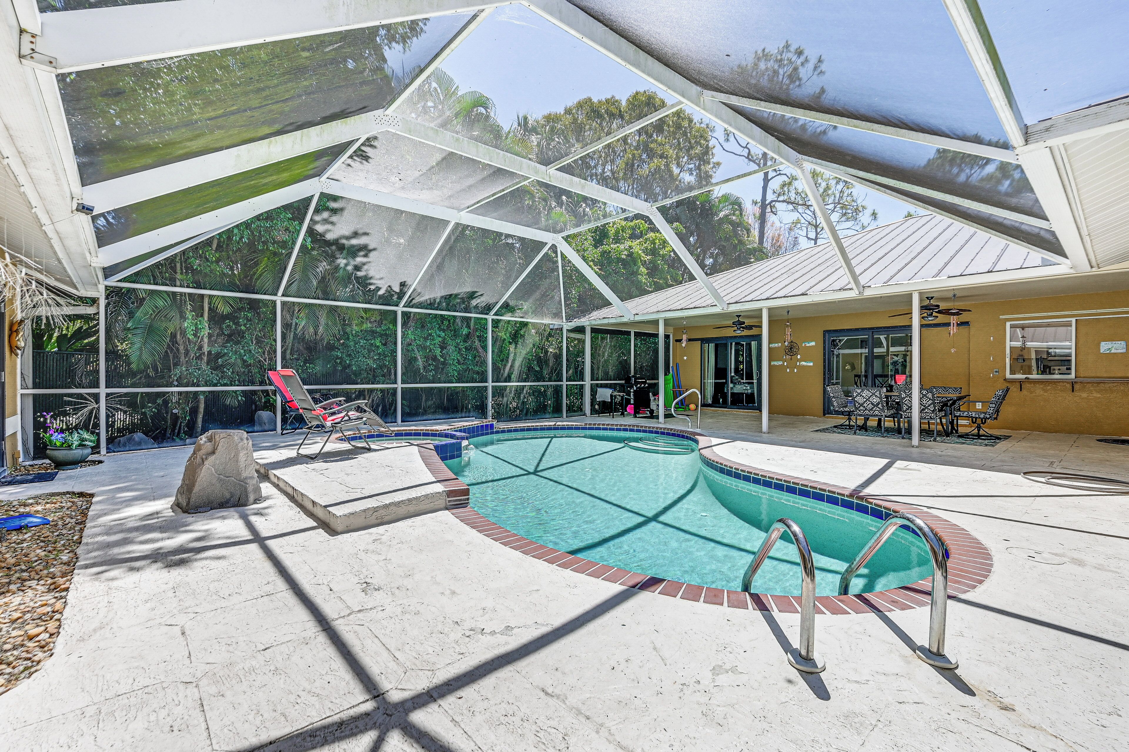 A covered outdoor swimming pool with a clear view of the surrounding area, including a patio with chairs and a table, and a building with a yellow exterior.