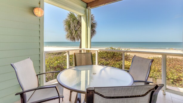 A covered outdoor seating area with a glass table and chairs, overlooking a beach.