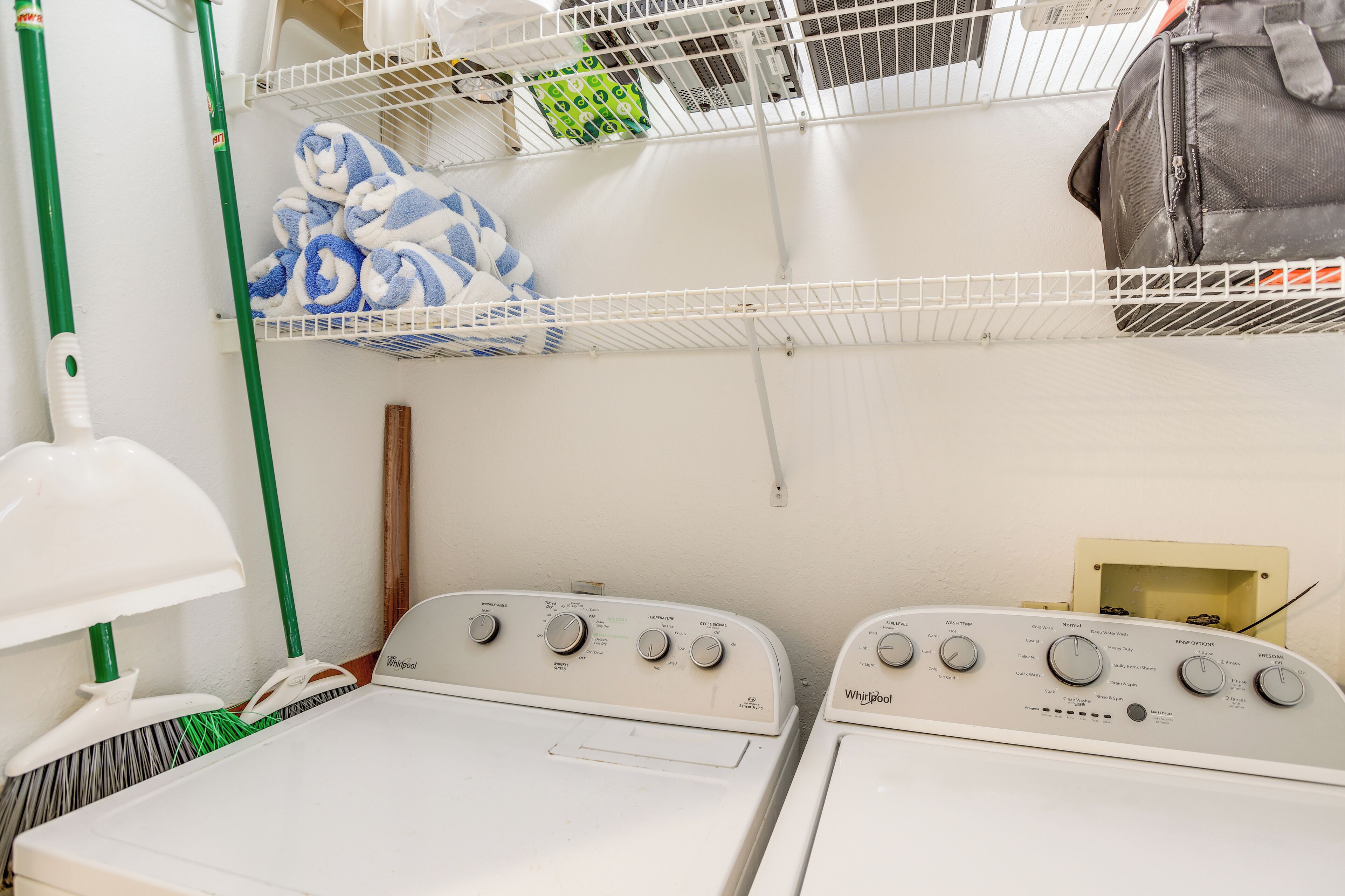 A laundry area with two Whirlpool washers, a green mop, a white floor lamp, and a shelf with towels and a bag.