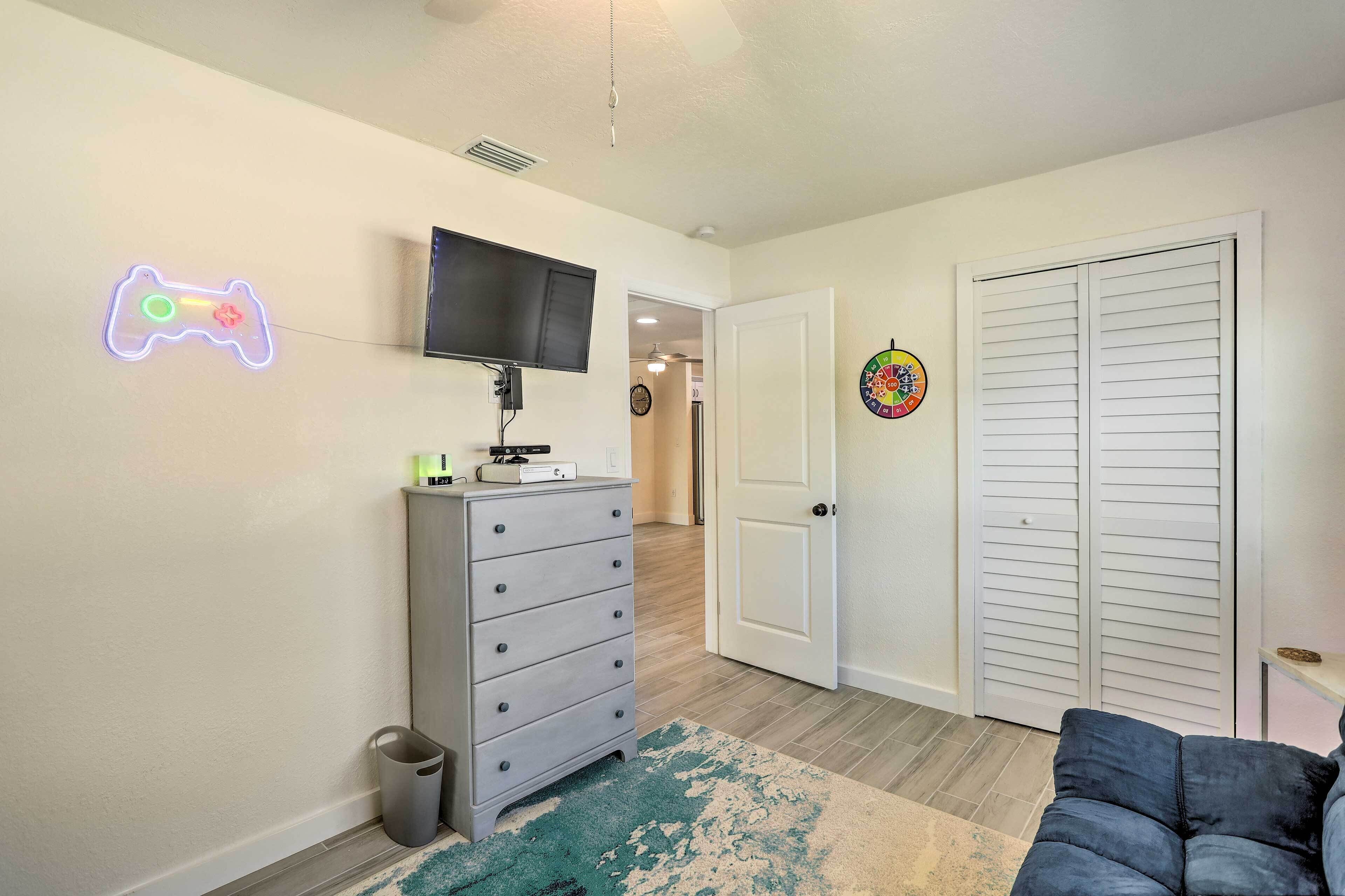 A room with a TV mounted on the wall, a neon game controller sign, a dresser, a closet, and a blue couch.