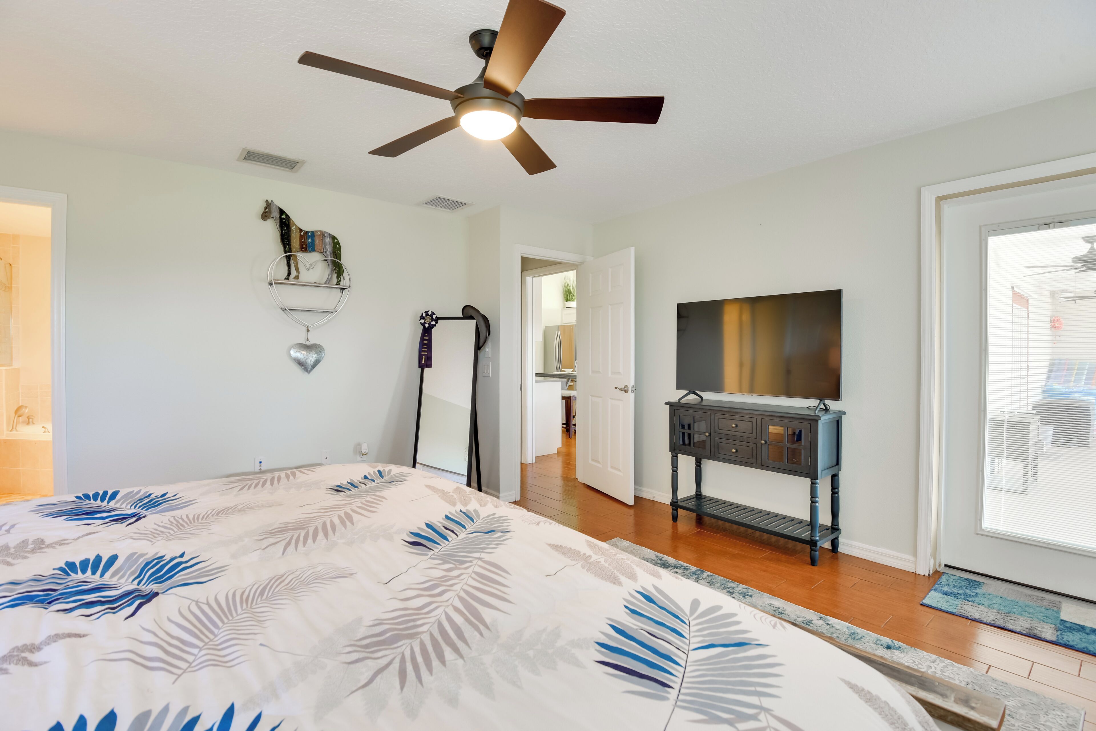 Un dormitorio con cama, ventilador de techo, televisor, cómoda y una puerta que da a otra habitación.