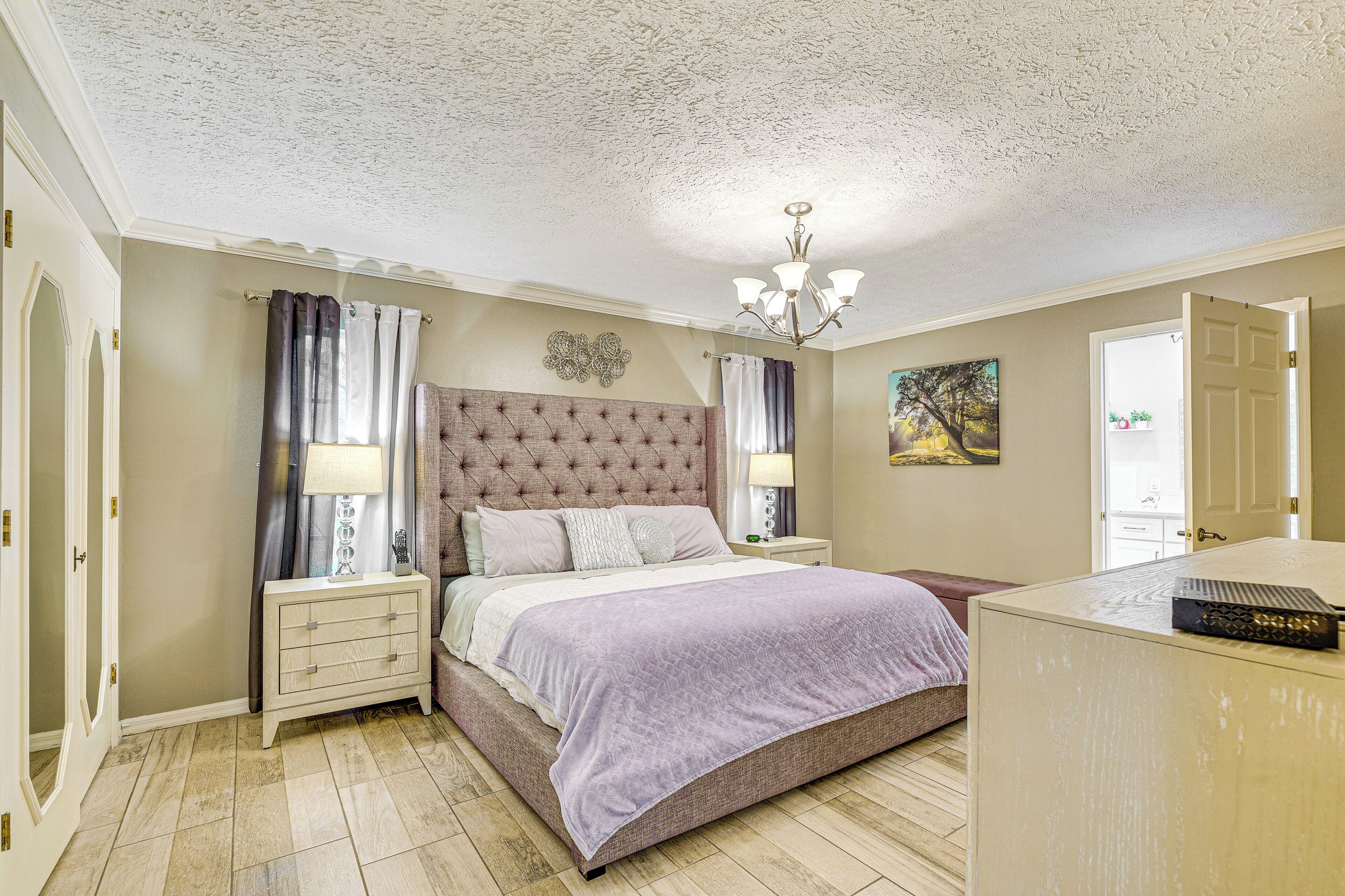 A bedroom with a tufted headboard, a bed with a purple bedspread, two bedside tables with lamps, a dresser, and a painting on the wall.