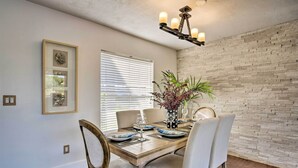 Un coin repas avec une table en bois, des chaises blanches et un lustre. On y trouve un mur en briques et une fenêtre avec des stores.