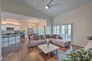Uma sala de estar ampla, com um sofá modular grande, uma mesa de centro e uma área de cozinha com armários brancos e micro-ondas.