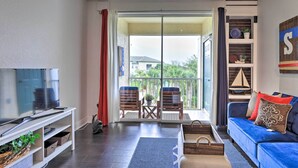 A living room with a TV, sofa, and chairs, a balcony with a view, and a wooden shelf with decorative items.