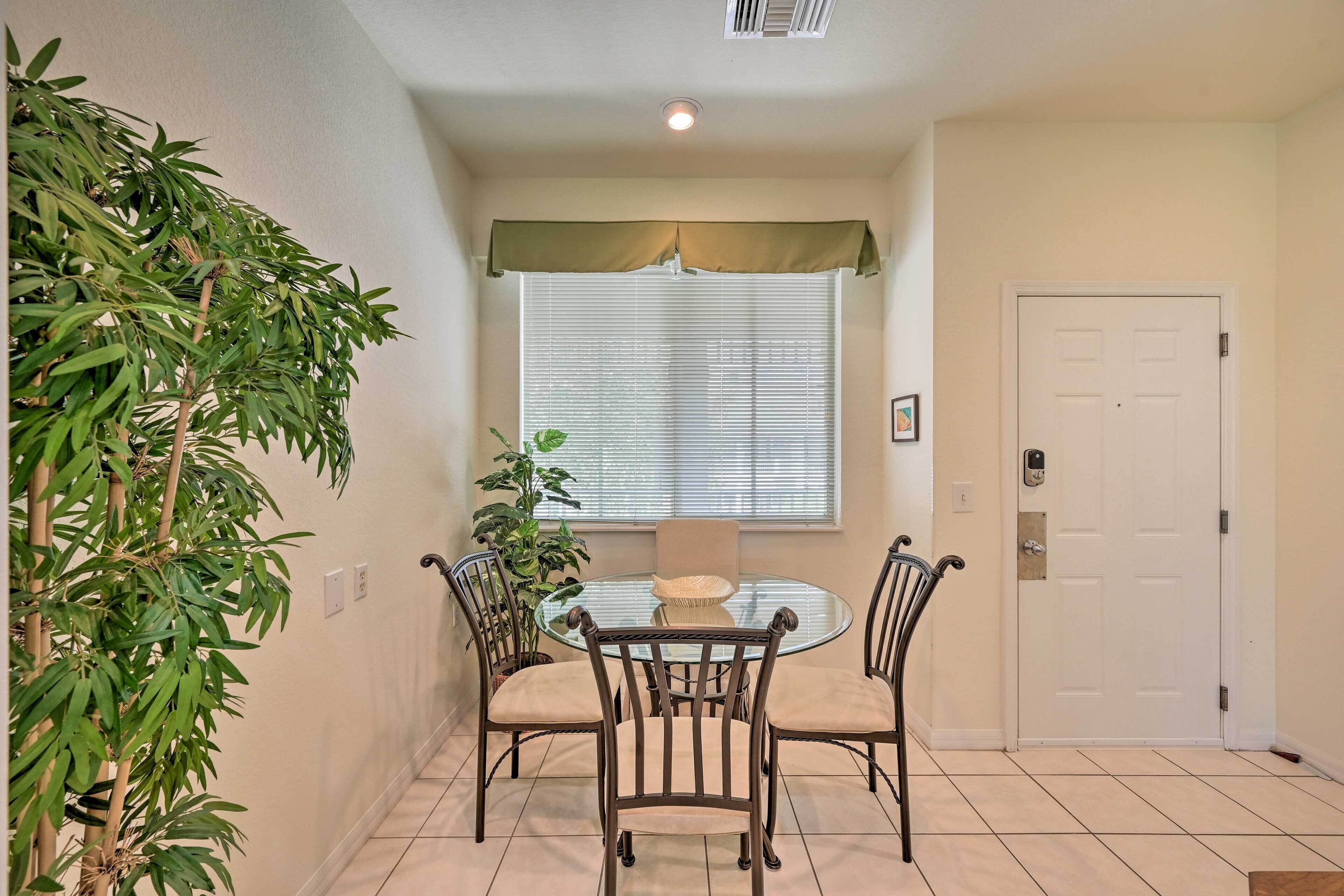 A dining area with a round glass table, four chairs, a potted plant, and a window with blinds.