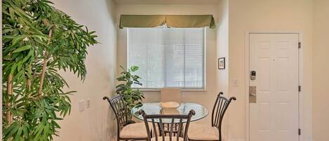 A dining area with a round glass table, four chairs, a potted plant, and a window with blinds.