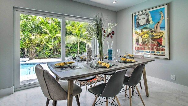 A dining area with a large table set for four, a view of a swimming pool, and a tropical garden visible through the glass doors. - Modern Wilton Manors Home w/ Outdoor Oasis! (Wilton Manors)