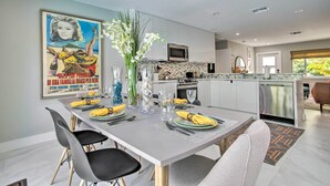 A modern dining area with a grey table set for four, surrounded by black chairs. A large poster adorns the wall, and a vase with white flowers is on the table. - Modern Wilton Manors Home w/ Outdoor Oasis! (Wilton Manors)