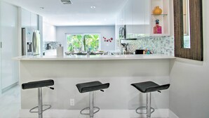 A modern kitchen with a central island, three bar stools, and a view of a dining area with a window. - Modern Wilton Manors Home w/ Outdoor Oasis! (Wilton Manors)