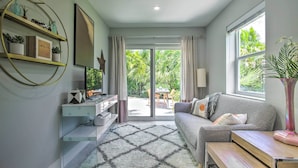 A modern living room with a sofa, a coffee table, and a TV on a stand. There is a large window with curtains, a door leading to a balcony, and a wall-mounted shelf with decorative items. - Modern Wilton Manors Home w/ Outdoor Oasis! (Wilton Manors)