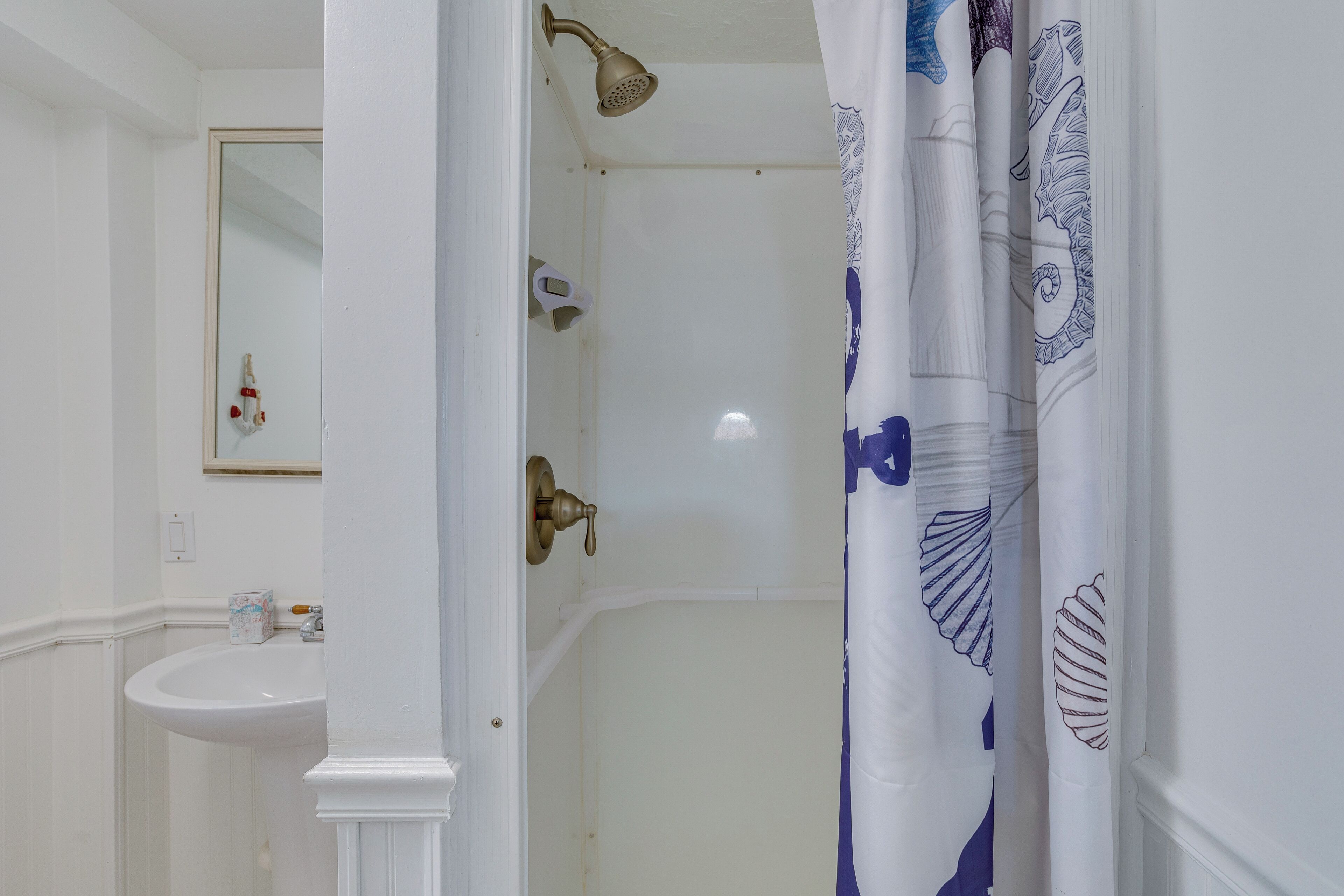 Un baño con lavamanos blanco, ducha con cabezal de ducha manual y cortina con estampado de hojas.