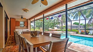 A poolside dining area with a wooden table and chairs, a ceiling fan, and a view of the pool and palm trees.