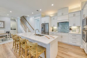 Une cuisine moderne avec des meubles blancs, un îlot central doté de tabourets, des appareils en acier inoxydable et un escalier en bois.