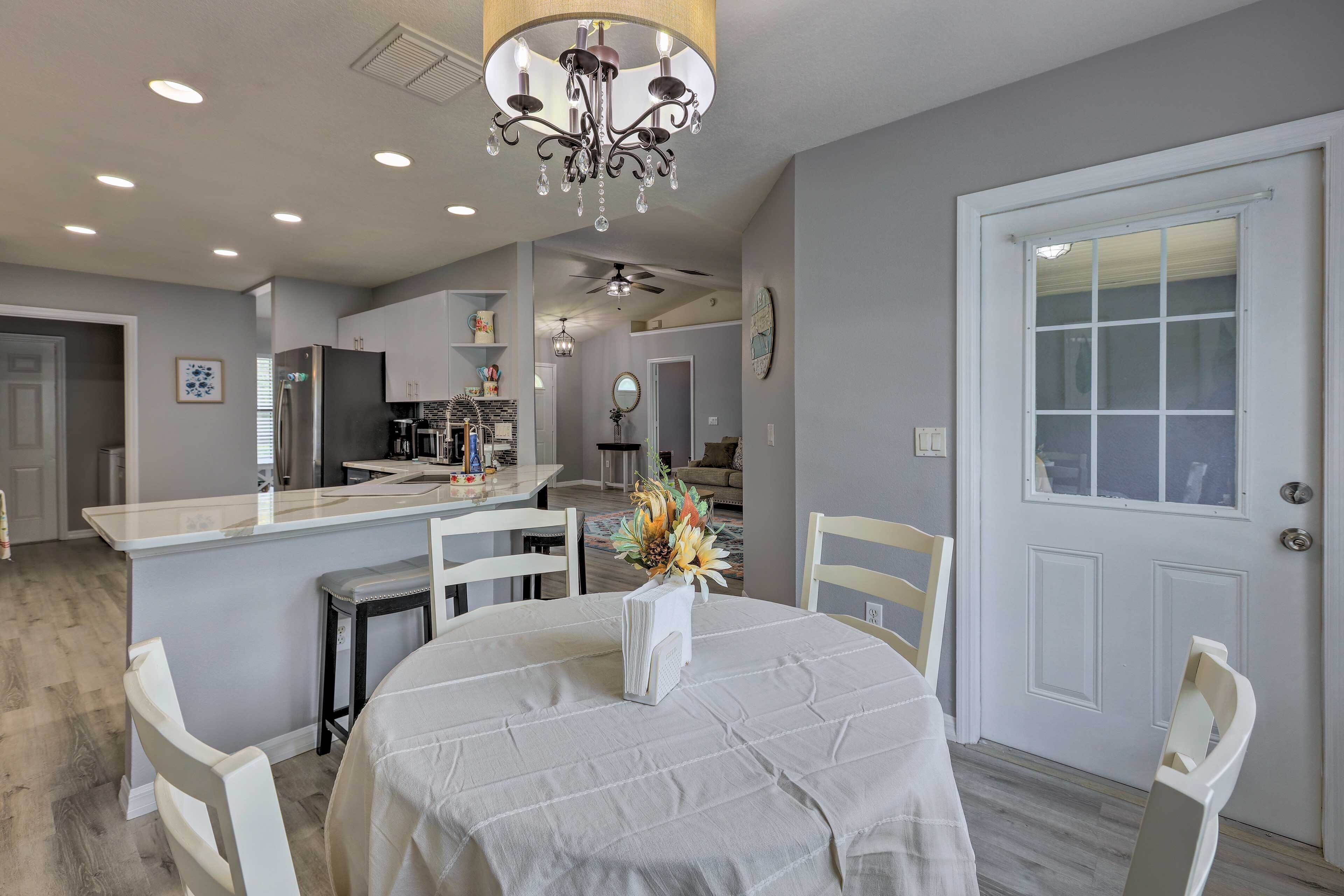 A dining area with a round table, white chairs, a chandelier, and a kitchen in the background.