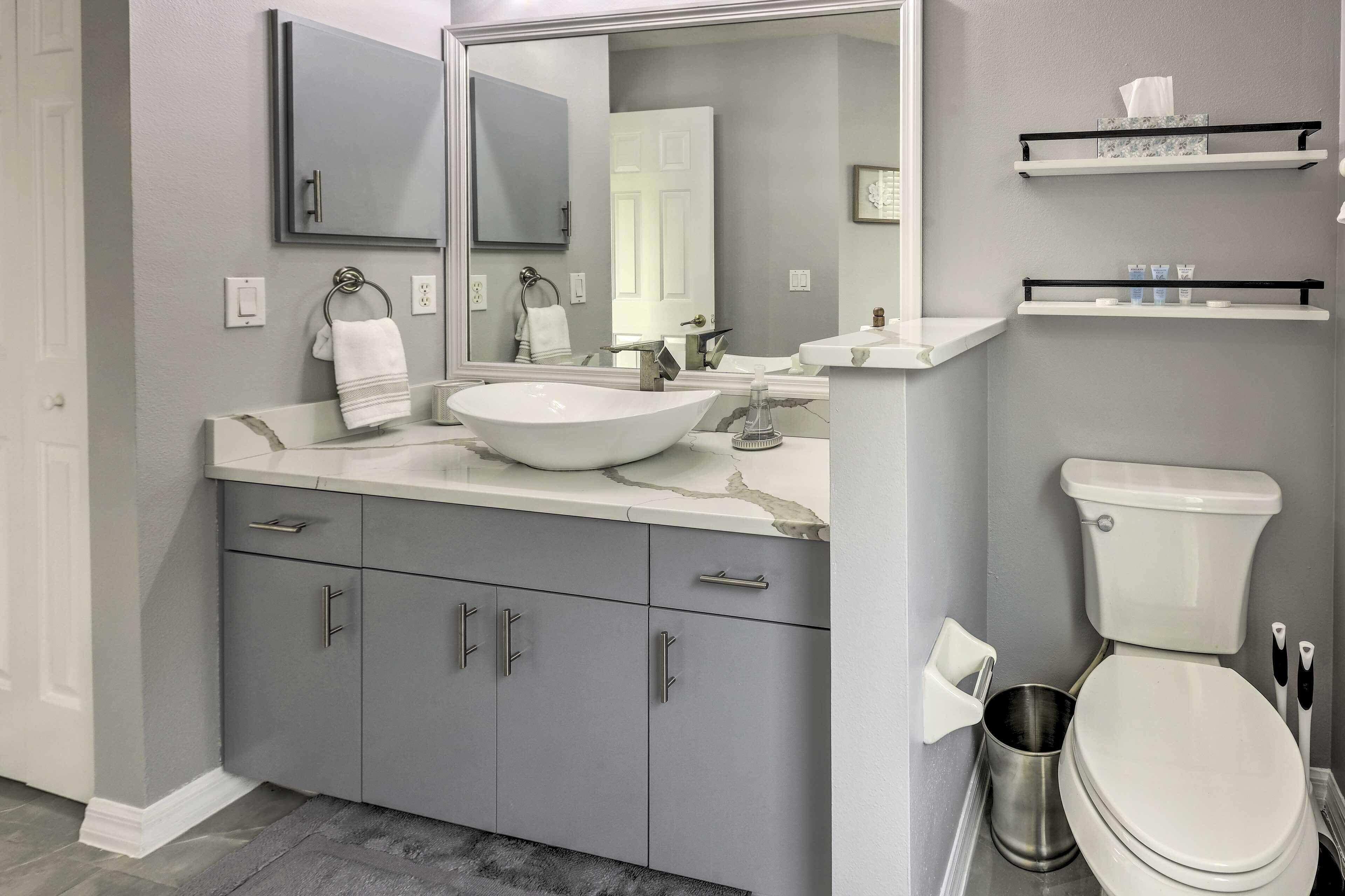 A modern bathroom with a large sink, mirror, and toilet. There are gray cabinets with silver handles and a small shelf above the toilet.