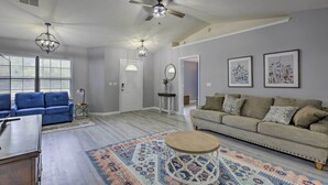 Una sala de estar con un sofá azul, un sofá color beige, una mesa de centro y un tapete estampado. Hay ventiladores de techo, una araña y obras de arte enmarcadas en las paredes.
