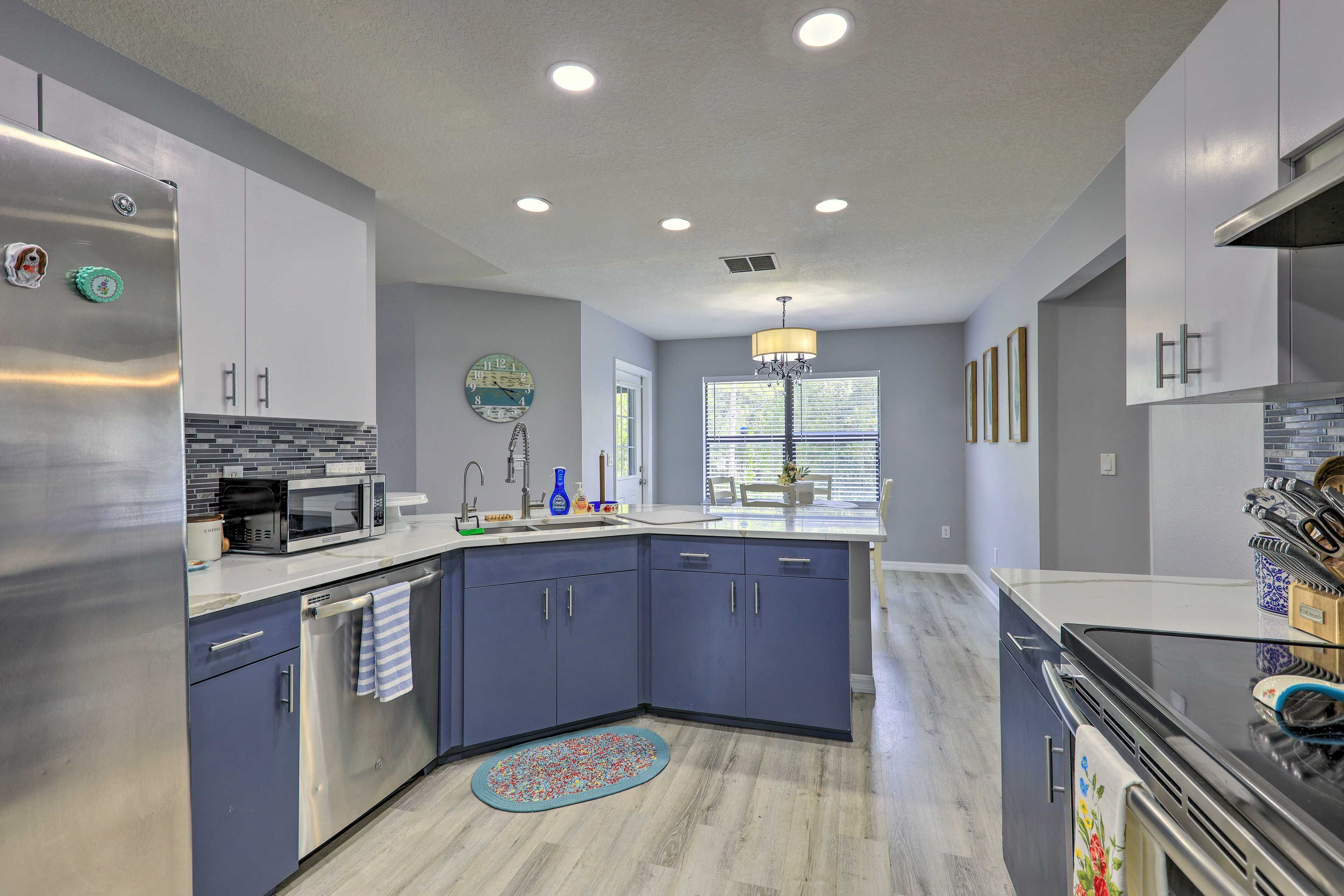 A modern kitchen with a stainless steel refrigerator, microwave, and dishwasher, a blue tiled backsplash, and a window with blinds.