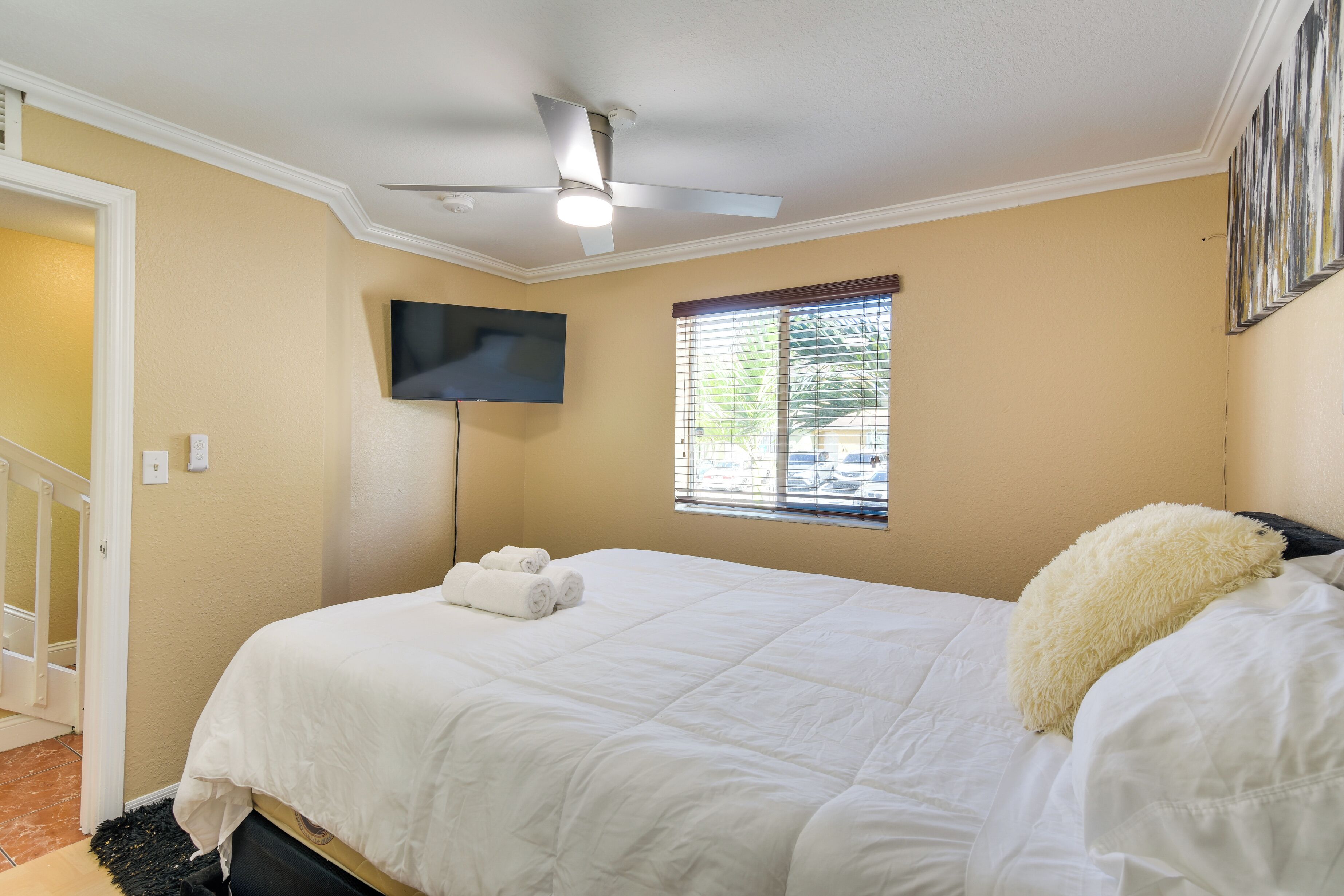 A bedroom with a bed, a ceiling fan, a TV mounted on the wall, and a window with blinds.
