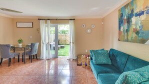 Una sala de estar con un sofá color turquesa, una mesa de comedor con sillas, un ventilador de techo y vistas al patio trasero a través de una puerta de vidrio.